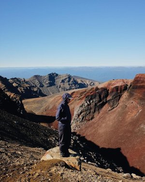 A person in a blue jacket stands on a rocky outcrop, overlooking a vast volcanic landscape with rugged mountains and clear skies.