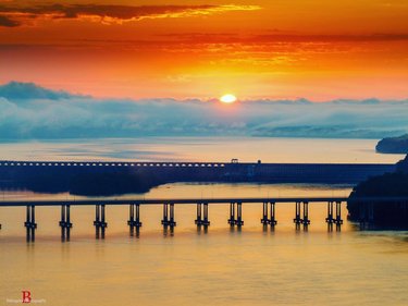 Sunset over a calm river, with a long bridge stretching across the water. Soft clouds reflect warm colors in the sky.