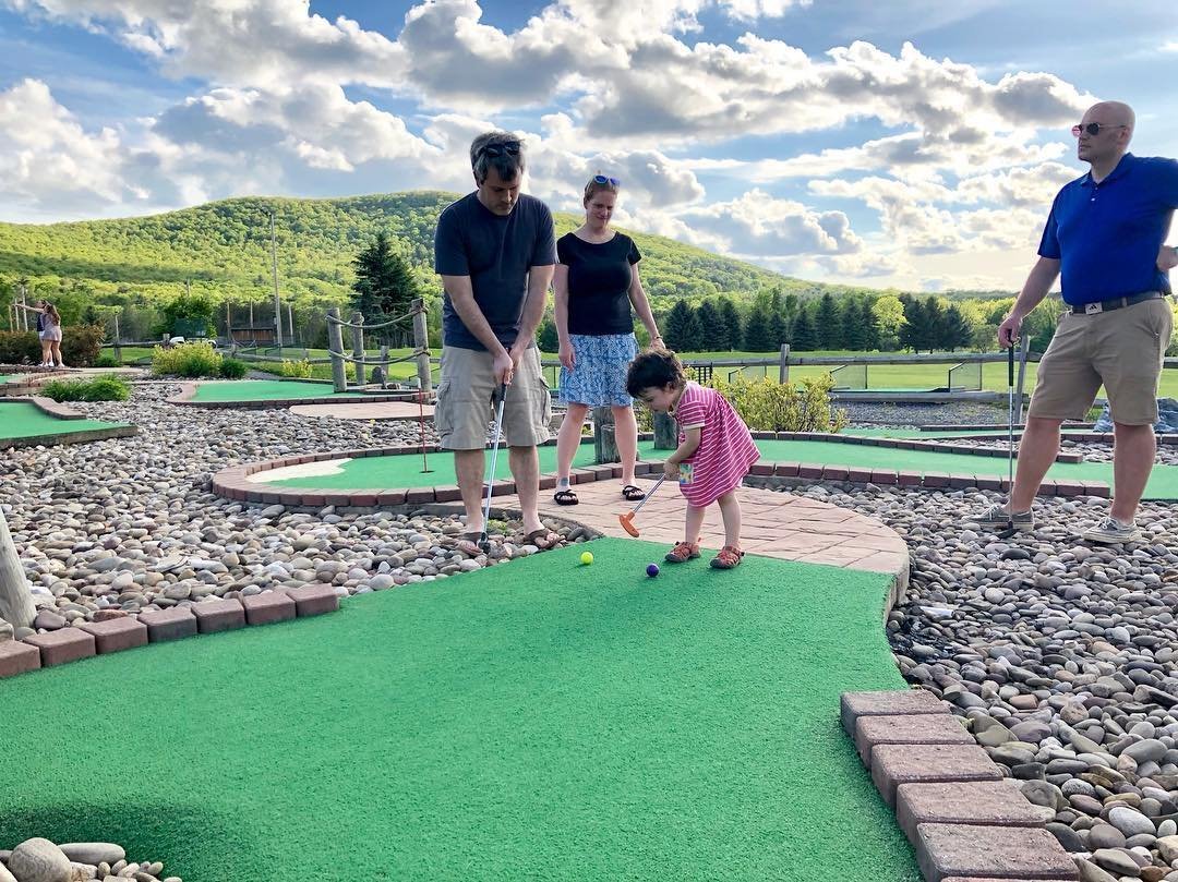 A family enjoys a game of mini-golf on a sunny day, surrounded by green hills and a blue sky with fluffy clouds. The scene captures a vibrant outdoor activity.