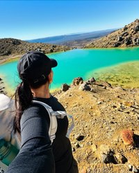 A hiker stands on rocky terrain, overlooking a vibrant turquoise lake surrounded by mountains under a clear blue sky.
