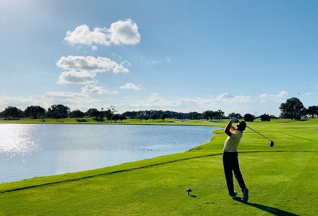 A golfer swings a club on a lush green course beside a shimmering lake, with blue skies and scattered clouds in the background.