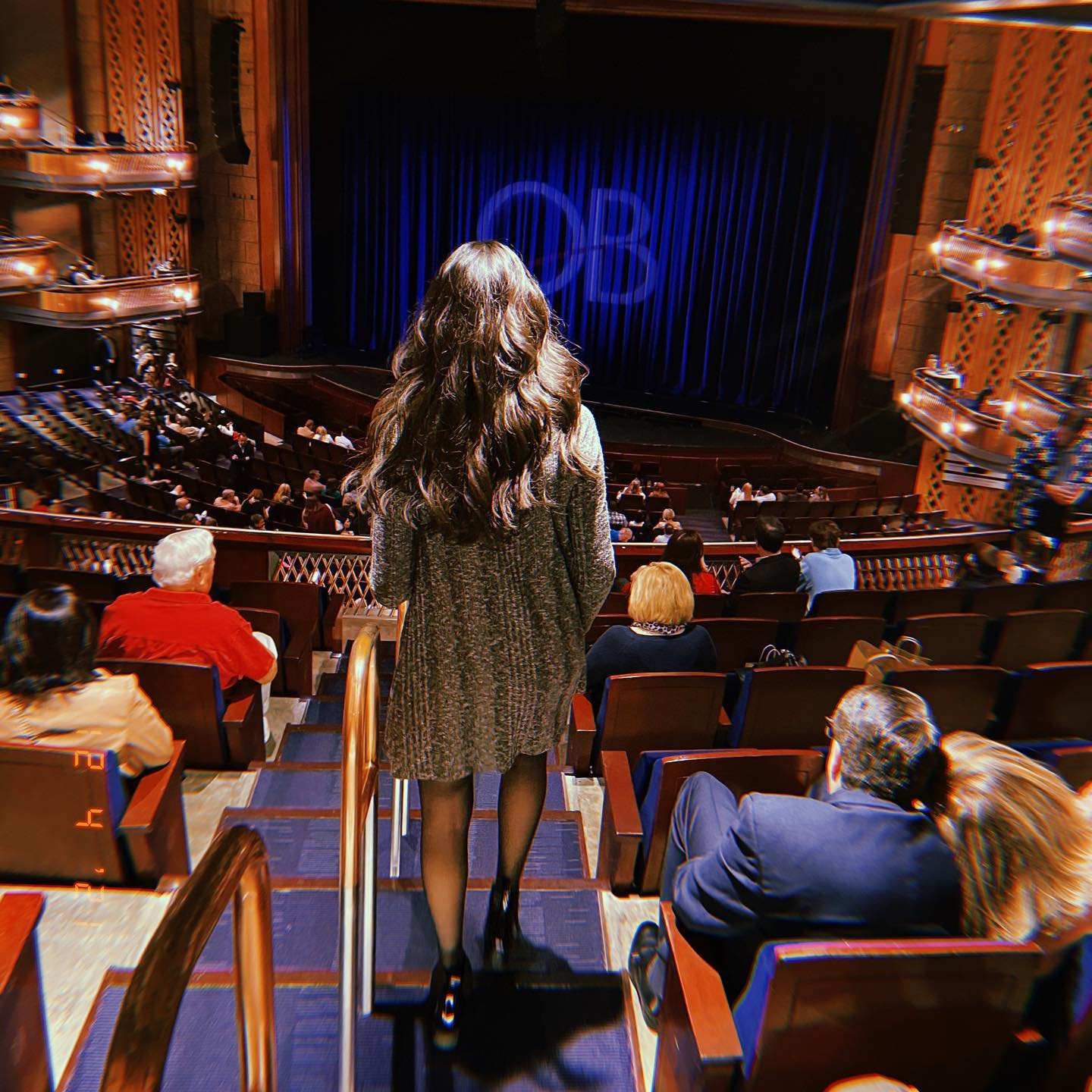 A woman with long hair walks down the stairs in a theater, facing a stage with blue curtains. Audience members are seated, awaiting a performance.