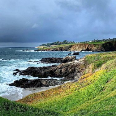 Coastal landscape featuring rocky shores and gentle waves under a cloudy sky. Lush green grass leads to the water's edge.