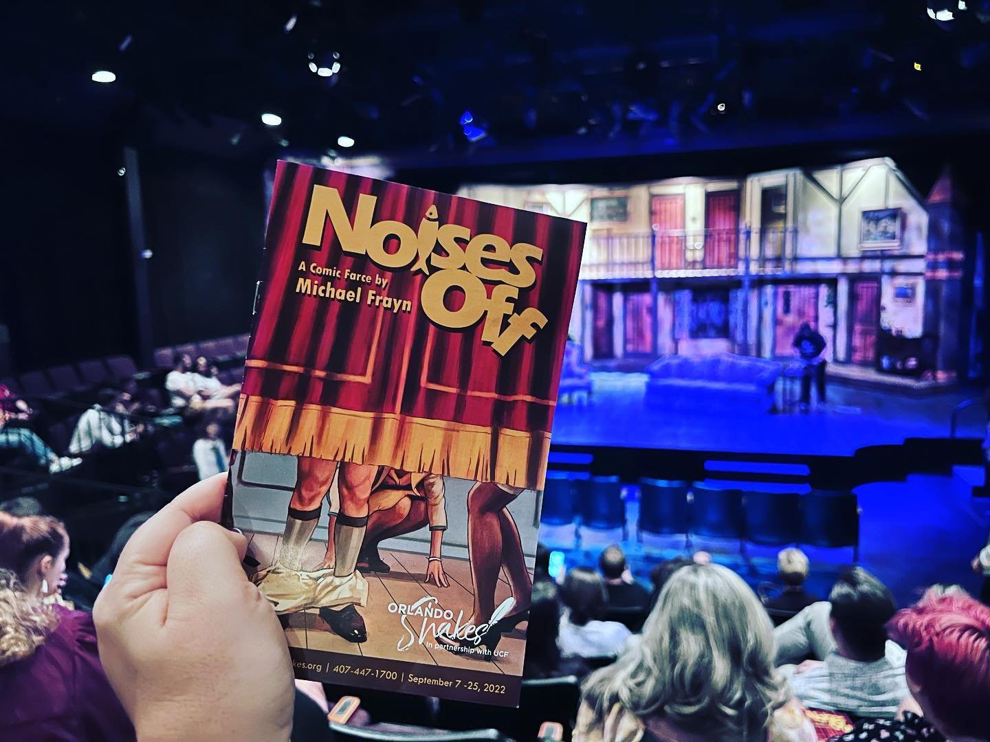 A hand holds a program for "Noises Off" in a theater. The stage is set with colorful backdrops, and an audience is visible in the background.