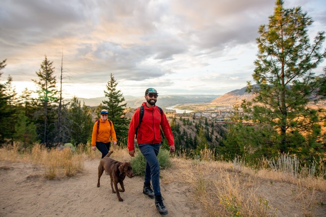 Two hikers, one in an orange shirt and the other in a red jacket, walk a brown dog along a sandy trail surrounded by trees. A scenic valley and mountains are visible in the background under a cloudy sky.