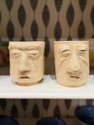 Two ceramic mugs with expressive, sculpted faces sit on a shelf, showcasing unique, artistic designs against a patterned background.