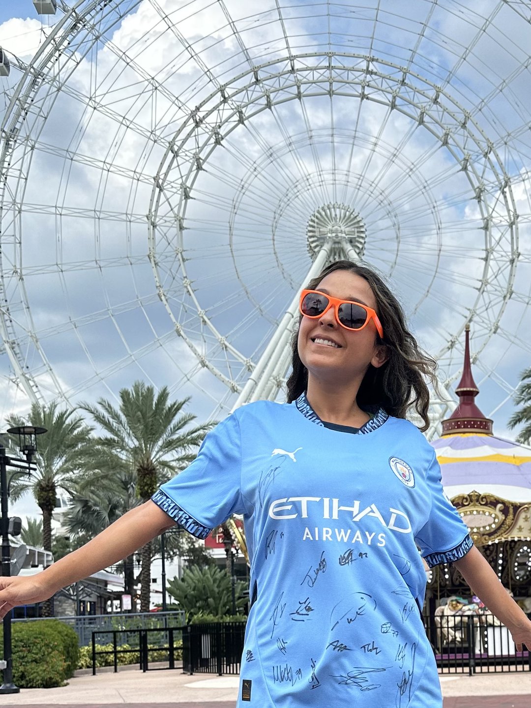 A young girl in a signed blue jersey and orange sunglasses smiles in front of a large Ferris wheel and palm trees, capturing a vibrant atmosphere.