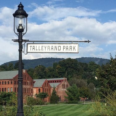 Signpost for Talleyrand Park with a vintage lamp, surrounded by green grass and historic brick buildings under a cloudy sky.