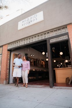 A couple stands outside a record store named "Hybrid Records," with shelves of vinyl visible inside. The storefront features modern lighting and a welcoming atmosphere.