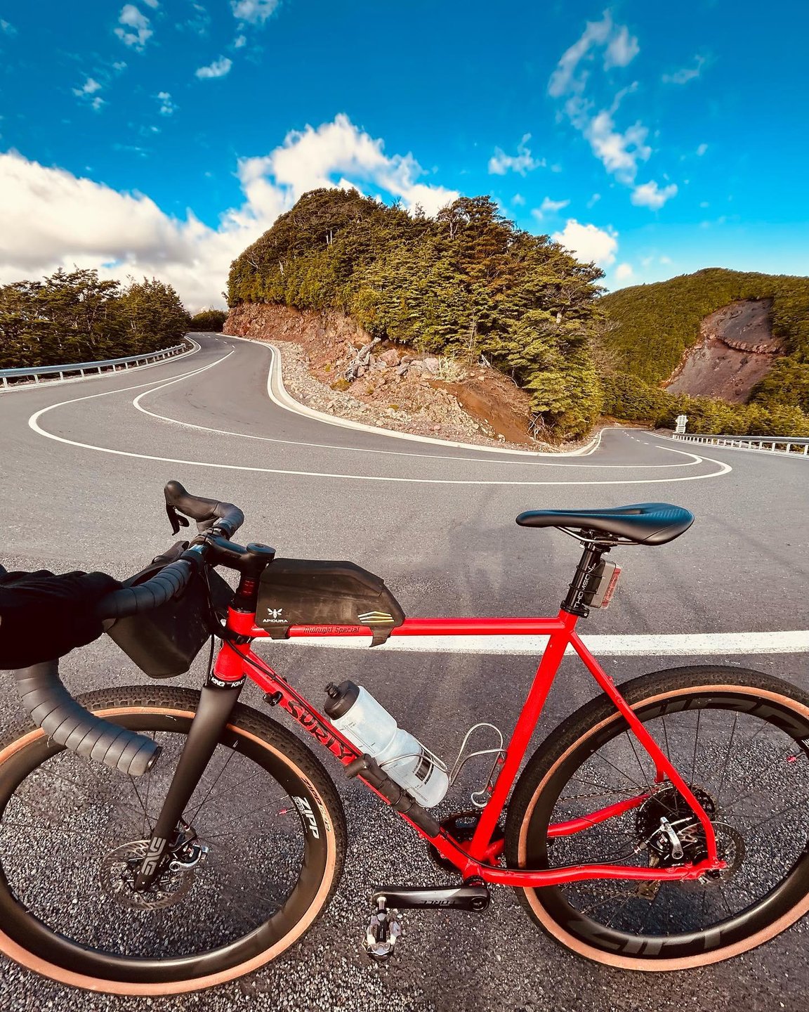 A red bicycle rests on a winding road surrounded by lush greenery and hills under a bright blue sky. The scene invites exploration and adventure.