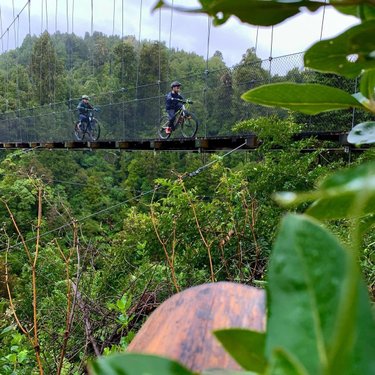 Two cyclists ride across a suspension bridge surrounded by lush greenery and mountains. The scene captures an adventurous outdoor experience.
