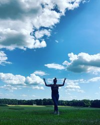 A person stands in a lush green field under a bright blue sky filled with fluffy white clouds, surrounded by trees in the distance.