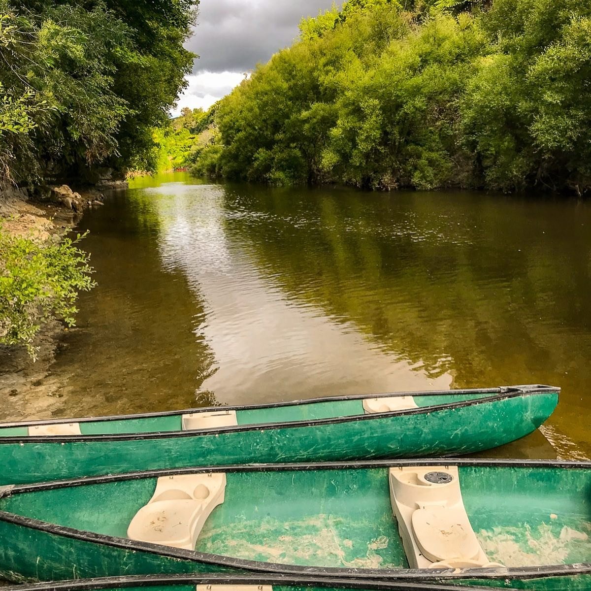 Two green canoes are moored at the edge of a calm river, surrounded by lush greenery and trees. The scene is tranquil, inviting exploration.