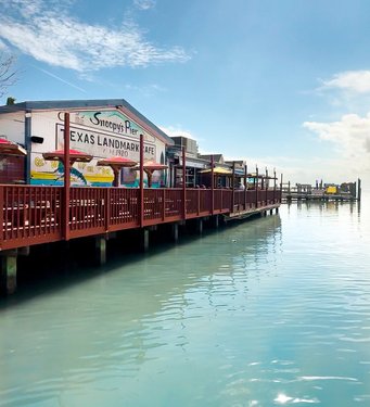 A wooden pier extends over calm, turquoise waters, featuring a colorful café with signage. Bright umbrellas line the deck under a clear blue sky.
