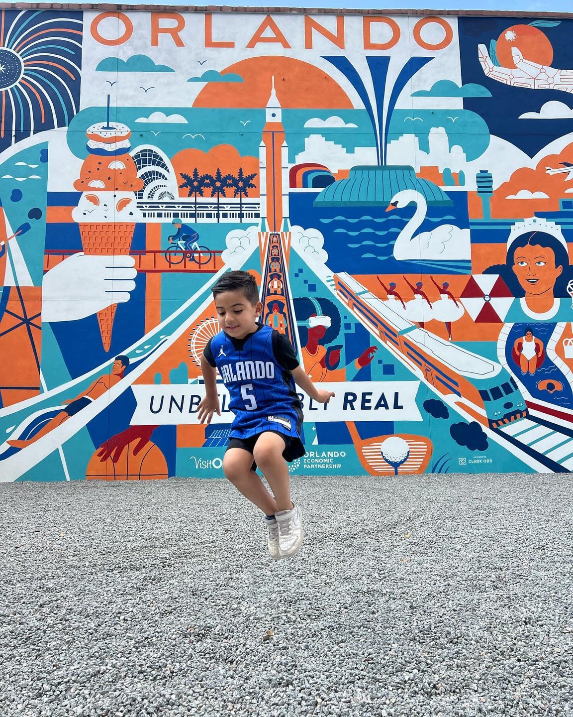 A child in a blue jersey jumps in front of a vibrant mural depicting Orlando's attractions, featuring ice cream, a swan, and iconic landmarks.