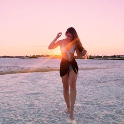 A woman walks barefoot on a sandy beach at sunset, with warm light illuminating her silhouette against a pastel sky.
