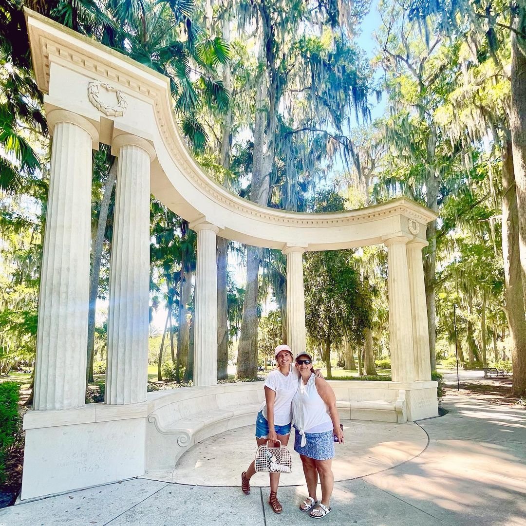 Two women stand under a grand, white-columned structure surrounded by lush greenery and hanging moss. Sunlight filters through the trees, creating a serene atmosphere.