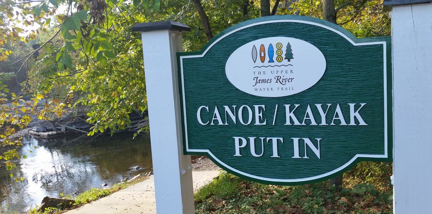 Sign for canoe and kayak put-in at the Upper James River Water Trail, surrounded by trees and a calm river. Ideal for outdoor activities.