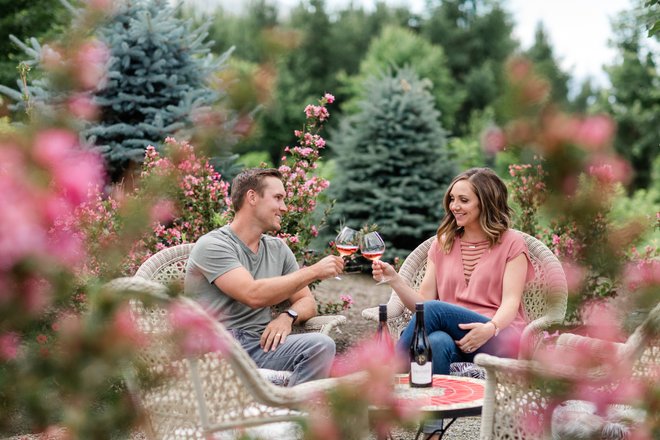 A couple toasts with wine glasses in a garden setting, surrounded by pink flowers and evergreen trees, seated in woven chairs.