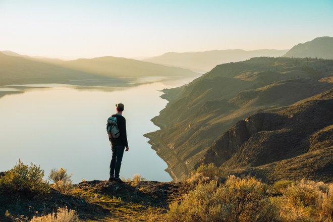 A hiker stands on a rocky outcrop overlooking a serene lake surrounded by rolling hills and mountains at sunset. The scene captures tranquility and natural beauty.