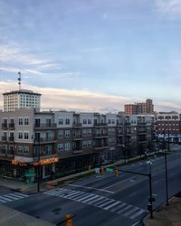 View of a city street featuring modern apartment buildings and a restaurant named "Newton Mushroom." The sky is partly cloudy, hinting at a vibrant urban atmosphere.