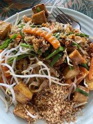 A plate of stir-fried noodles topped with vegetables, tofu, and crushed peanuts, set against a tropical leaf background.