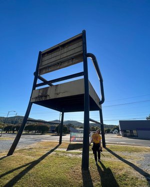 A person stands beside a giant metal chair in a grassy area, with mountains in the background and a clear blue sky above.