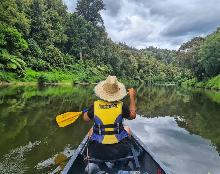 A person in a straw hat paddles a canoe on a calm river, surrounded by lush green trees and a cloudy sky, reflecting on the water.
