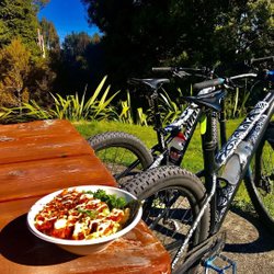 A bowl of food sits on a wooden table beside two mountain bikes, surrounded by lush greenery and trees in a sunny outdoor setting.
