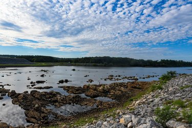 A serene river landscape with rocky shores, calm water reflecting a blue sky dotted with clouds, and lush greenery in the background.