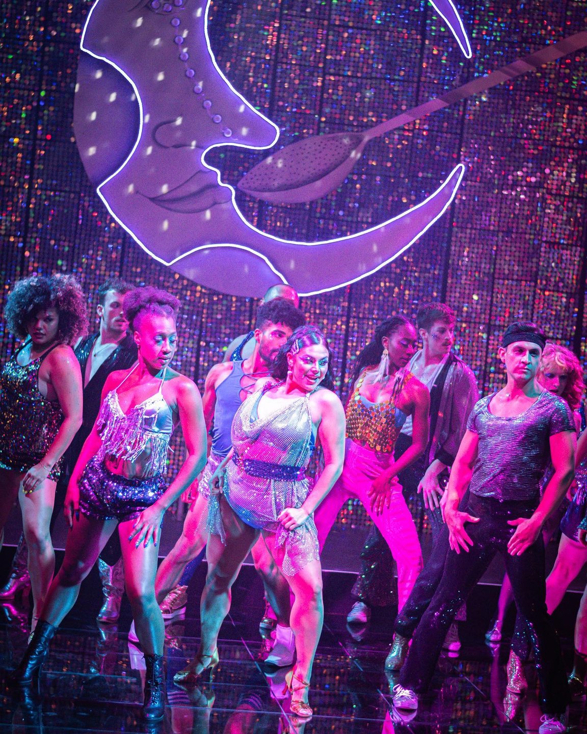 A vibrant stage performance featuring a diverse group of dancers in sparkling costumes, illuminated by colorful lights and a large moon backdrop.