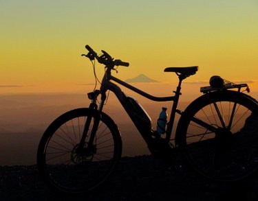 Silhouette of a bicycle against a sunset backdrop, with distant mountains visible. A water bottle is attached to the bike frame.