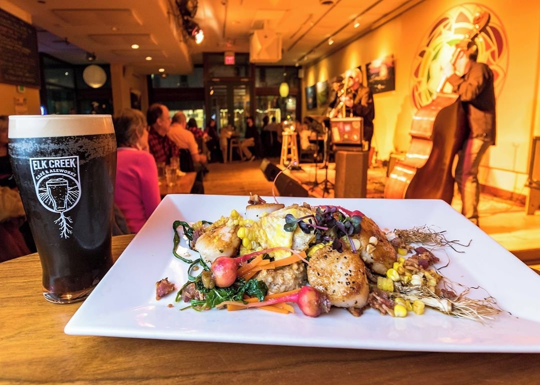 A plate of vibrant seafood and vegetables sits on a bar, with a dark beer beside it. A live band performs in a cozy restaurant setting.