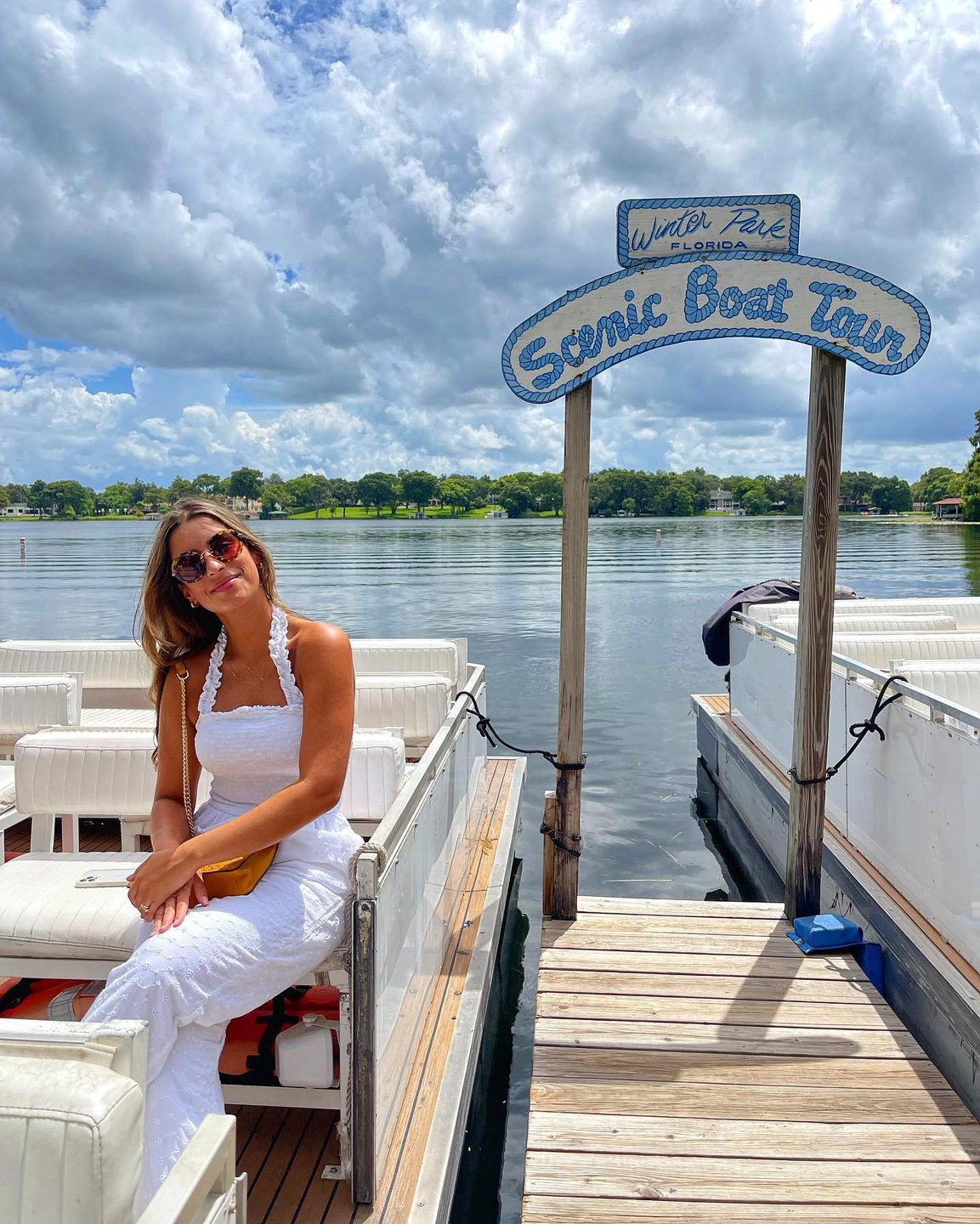 A woman sits on a boat dock next to a sign reading "Scenic Boat Tour," with calm waters and lush greenery in the background.