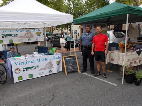 A vibrant market scene featuring a police officer and a cyclist beside vendor tents. Bicycles and local goods are displayed, inviting exploration.