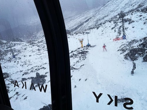 View from a gondola showing a snowy landscape with skiers and snowboarders on a slope, surrounded by rocky terrain and cloudy skies.