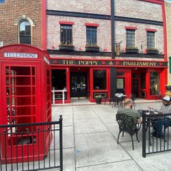 A vibrant red telephone booth stands beside a lively pub, "The Poppy & Parliament," with outdoor seating and brick architecture.