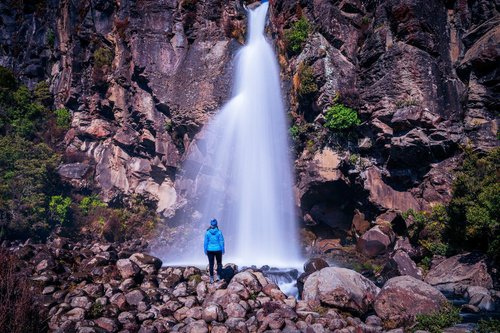 A person in a blue jacket stands on rocks near a cascading waterfall, surrounded by lush greenery and rugged cliffs. The scene captures natural beauty and adventure.