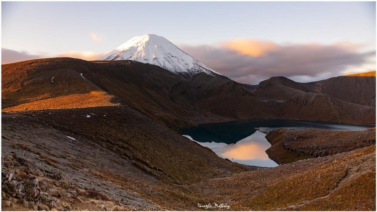 Snow-capped mountain rises above a serene lake, surrounded by rugged terrain and rolling hills under a cloudy sky. Ideal for nature exploration.