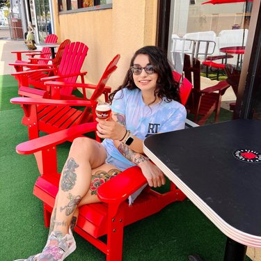 A young woman sits in a red Adirondack chair, enjoying ice cream outside a café. Brightly colored chairs and a green turf surface create a vibrant atmosphere.