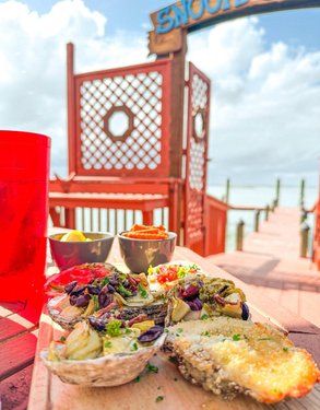 A wooden platter with baked oysters topped with herbs and sauces, set on a dock with a sign reading "Snoopy's" in the background.