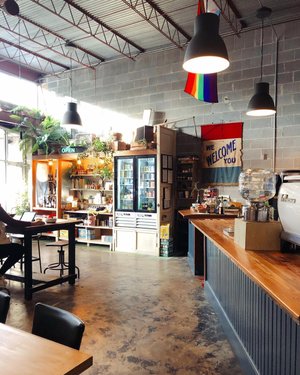 Cozy café interior featuring wooden counters, shelves with local goods, and a welcoming sign. Bright plants and colorful flags enhance the atmosphere.
