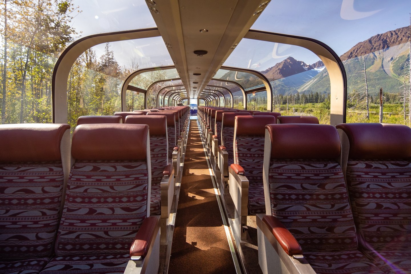 Interior view of a scenic train car with rows of plush seats, large windows showcasing mountains and greenery outside.