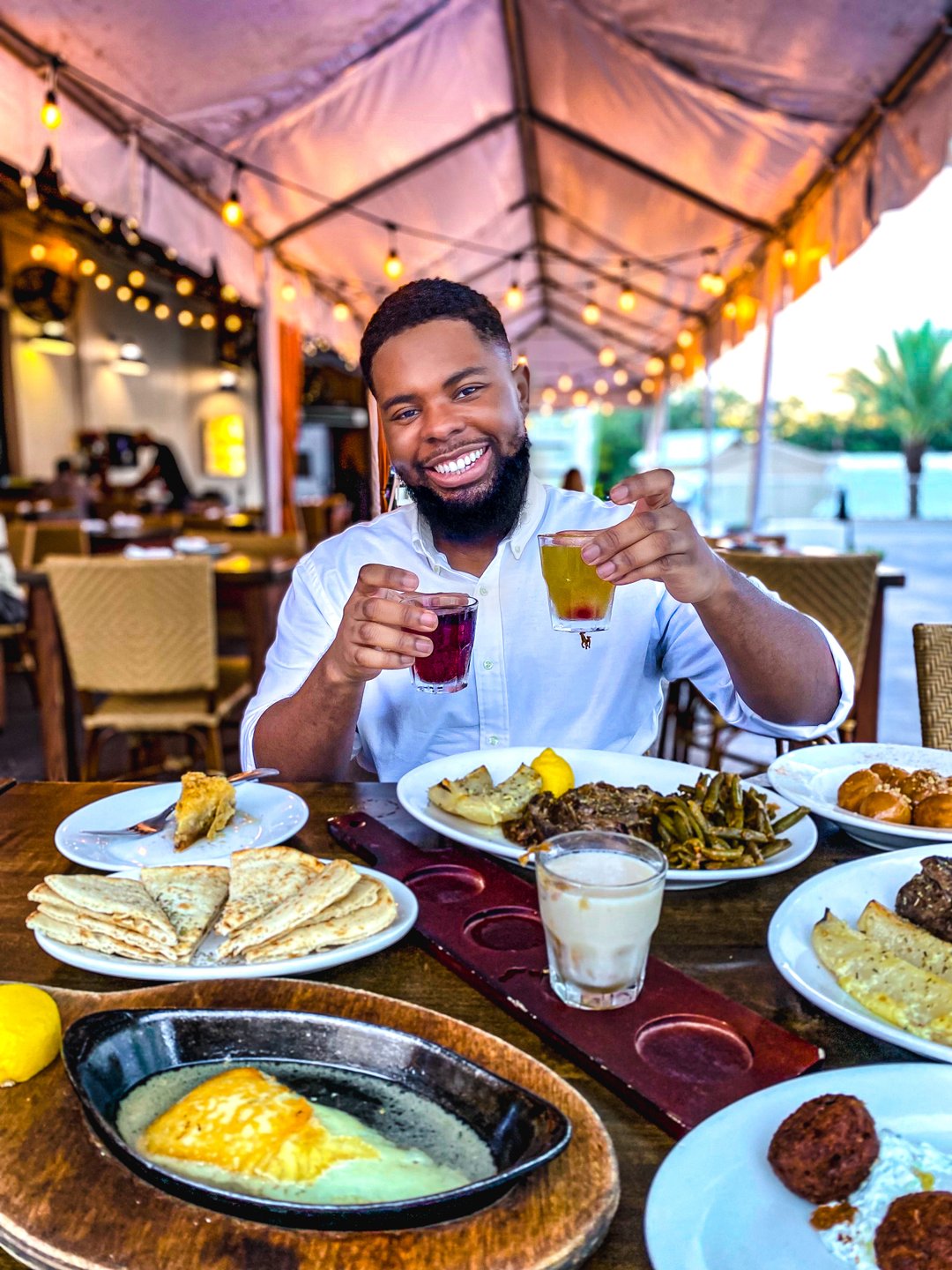 A man smiles while holding two colorful drinks at a table filled with various dishes, under a tented outdoor dining area.