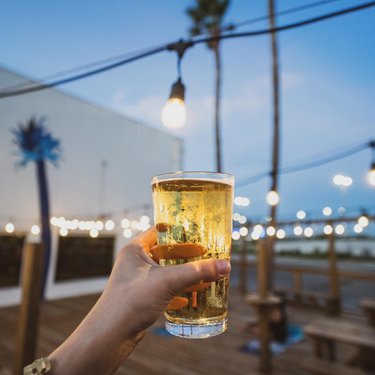 A hand holds a glass of golden beer against a backdrop of string lights and a twilight sky, creating a relaxed outdoor atmosphere.