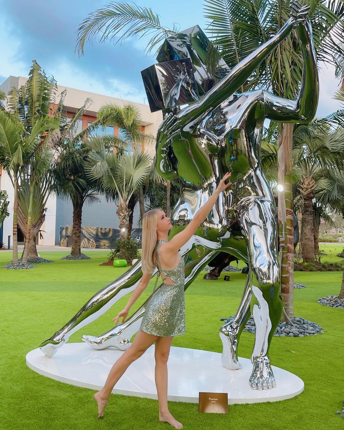 A woman in a shimmering dress poses beside a large, reflective sculpture of two figures in a lush, green garden setting. Palm trees and modern architecture are visible in the background.