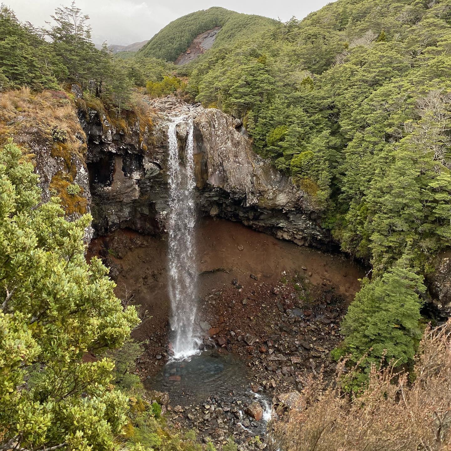 A cascading waterfall flows into a rocky pool, surrounded by lush green trees and rugged terrain, set against a mountainous backdrop.