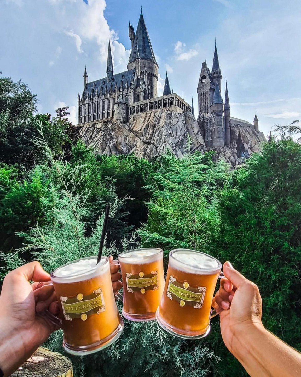 Three frosty mugs of butterbeer are held up in front of a grand castle surrounded by lush greenery under a bright sky.