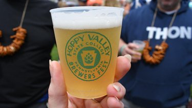 A hand holds a clear cup of beer with a green logo for Hoppy Valley Brewers Fest. People in the background enjoy the festival atmosphere.