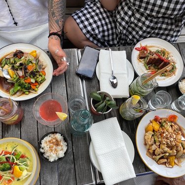 A wooden table displays an array of colorful dishes, including seafood, stir-fried vegetables, and salads, alongside drinks and utensils.